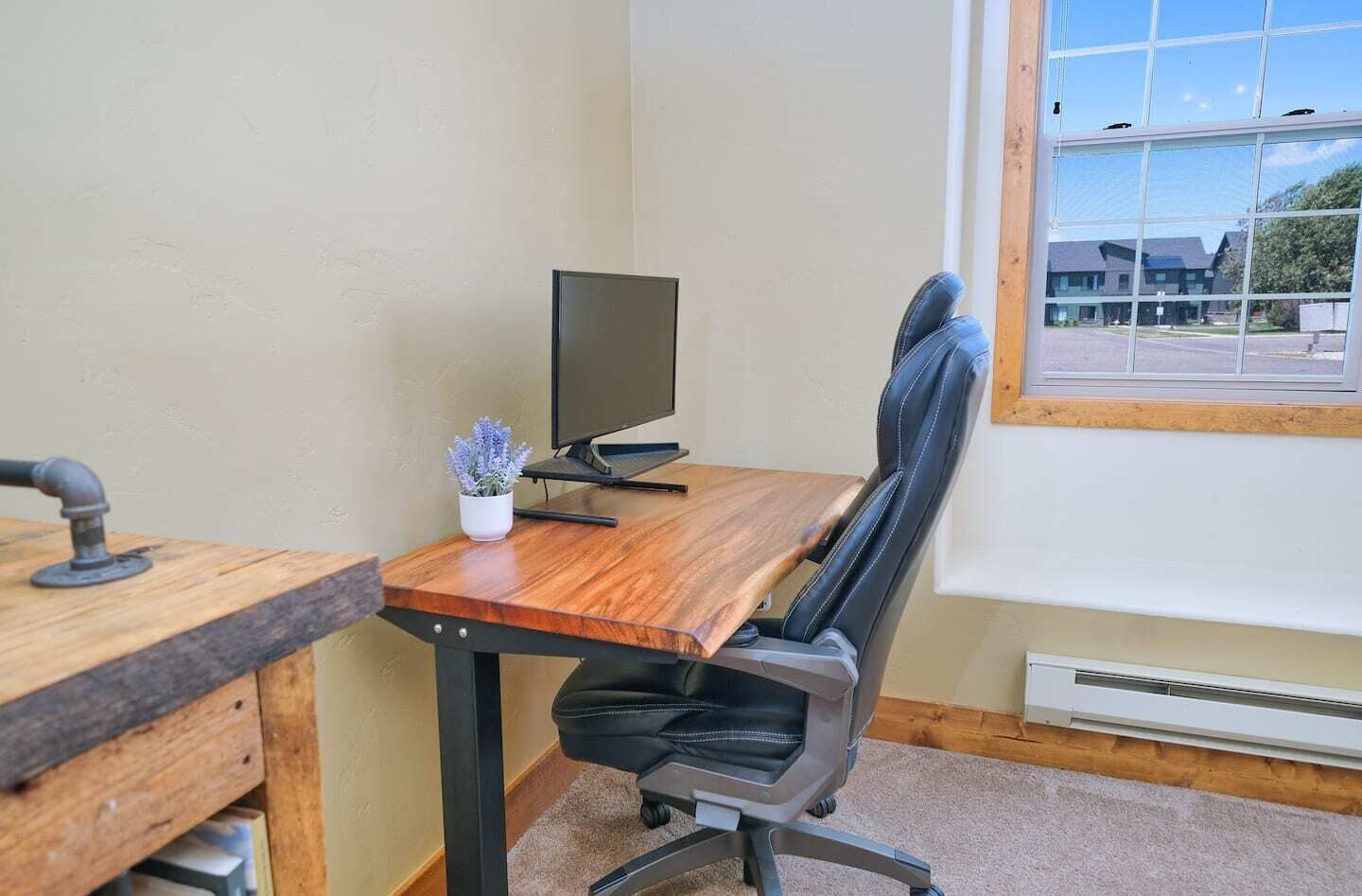 Un petit bureau avec un bureau en bois, une chaise de bureau noire, un écran d’ordinateur et une fenêtre donnant sur des immeubles.