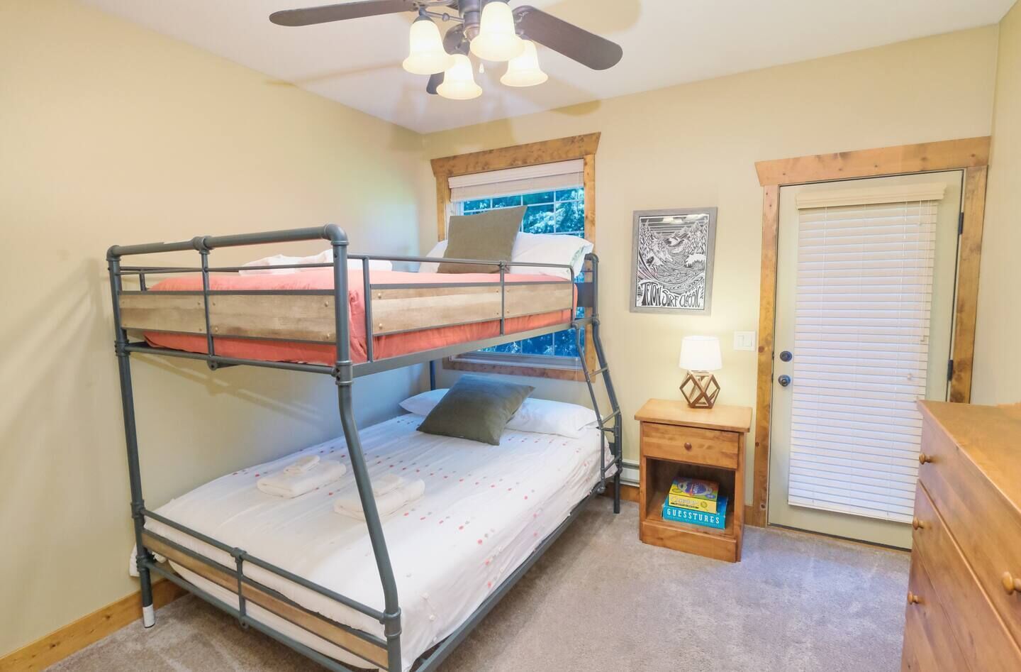 Habitación con literas, ventilador de techo, ventana con vistas, puerta y una mesita de noche pequeña con lámpara y una caja etiquetada 'GUESTSTURES'.