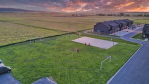 Una zona residencial con un amplio campo abierto, un espacio pavimentado con algunas personas y una hilera de casas con entrada para vehículos.