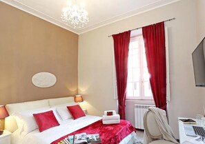A hotel room with a bed, a desk, a chair, a radiator, and a window with red curtains. - Liberty Rome Apartment (Roma)