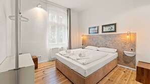 A modern bedroom with a wooden floor, a large bed with white linens, a window with sheer curtains, and two wall-mounted light fixtures. - Boxi 6 pax - Roomy Apartment with balcony (Berlin)