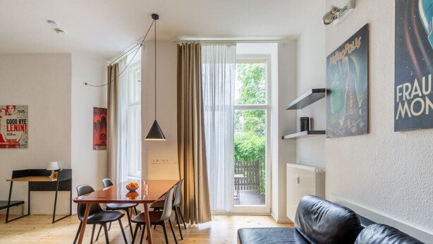 A modern living room with a wooden floor, a black leather sofa, a dining table with chairs, and a large window with curtains. - Boxi Beautifull Studio with patio (Berlin)
