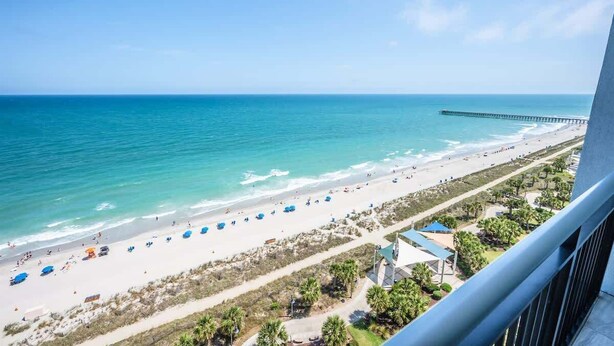 A beach view from a balcony with a pier extending into the ocean, beach umbrellas, and a clear sky. - Direct View Centrally located Oceanfront Boardwalk (Myrtle Beach)
