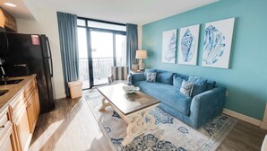 A living room with a blue sofa, a coffee table, and a large window with curtains. - Newest at Beach Cove,Updated Ocean Front,Low Floor (North Myrtle Beach)