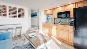 A modern kitchen with wooden cabinets, a black refrigerator, and a microwave. There is a dining area with a blue sofa and a patterned rug. - Newest at Beach Cove,Updated Ocean Front,Low Floor (North Myrtle Beach)