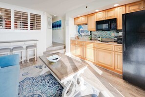 A modern kitchen with wooden cabinets, a black refrigerator, and a microwave. There is a dining area with a blue sofa and a patterned rug. - Newest at Beach Cove,Updated Ocean Front,Low Floor (North Myrtle Beach)