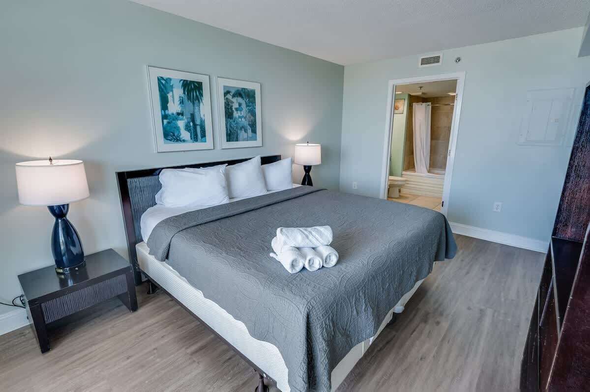 A neatly made bed with a grey blanket and white pillows, two bedside lamps, and two framed artworks on the wall.