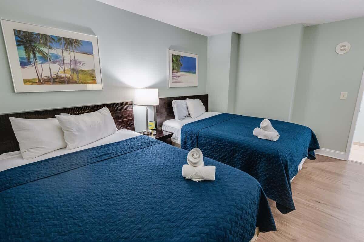 A hotel room with two beds, each with a blue bedspread and white pillows. There are two framed pictures on the wall, one depicting a beach scene and the other a tropical landscape.