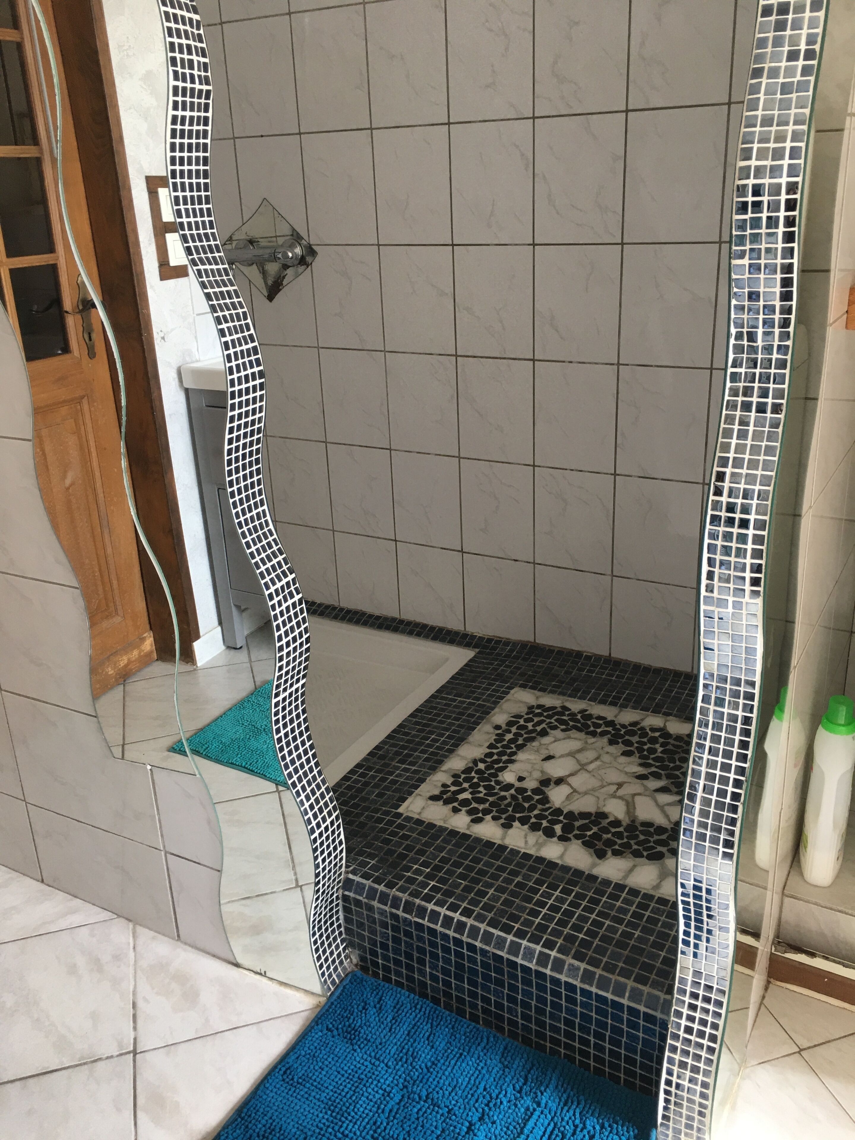 Un bany amb una zona de dutxa revestida amb mosaic blanc i negre, un tapet blau i una porta de fusta.