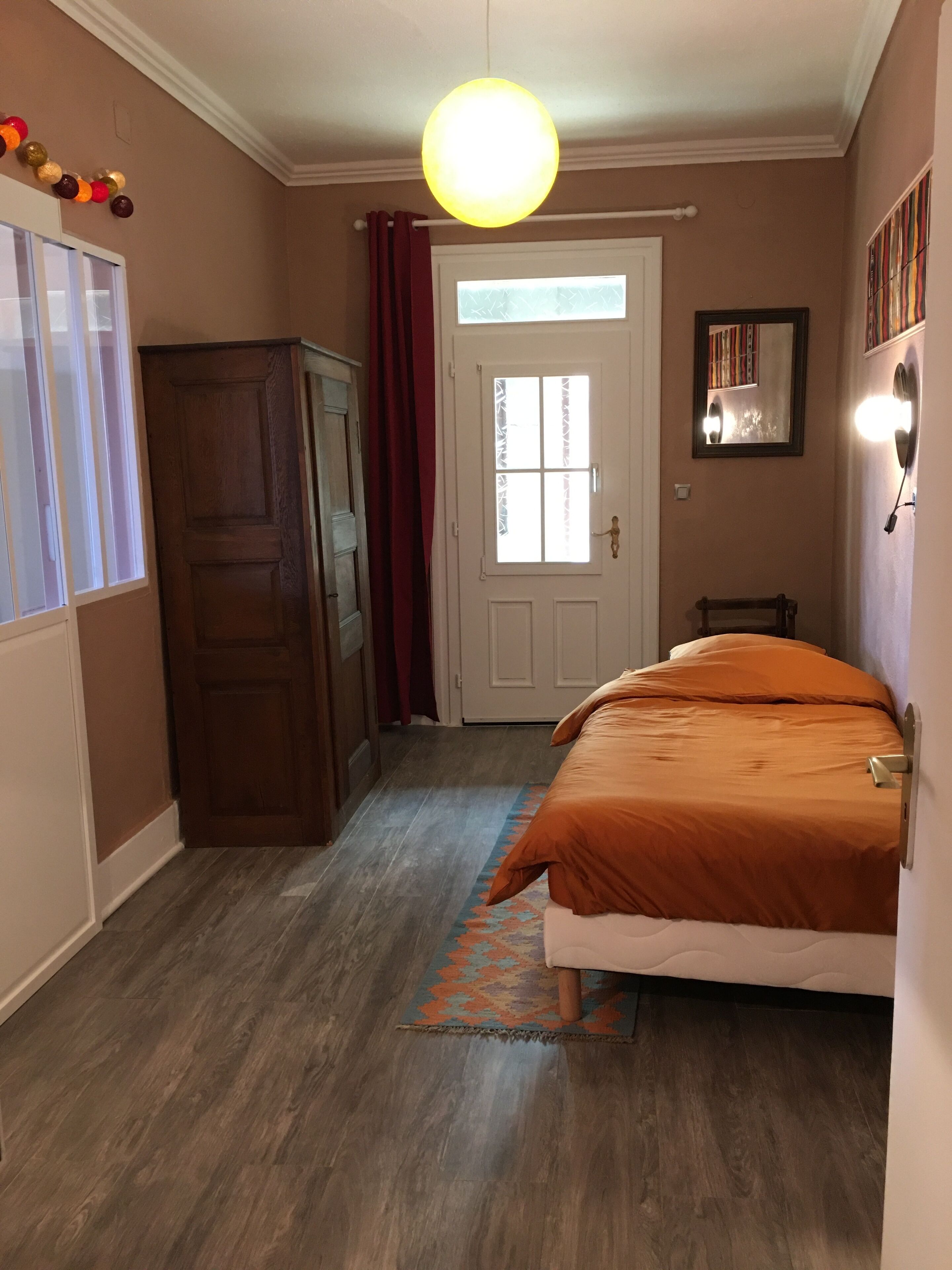 Un dormitorio con cama, armario, puerta con cortinas, espejo y lámpara colgante.