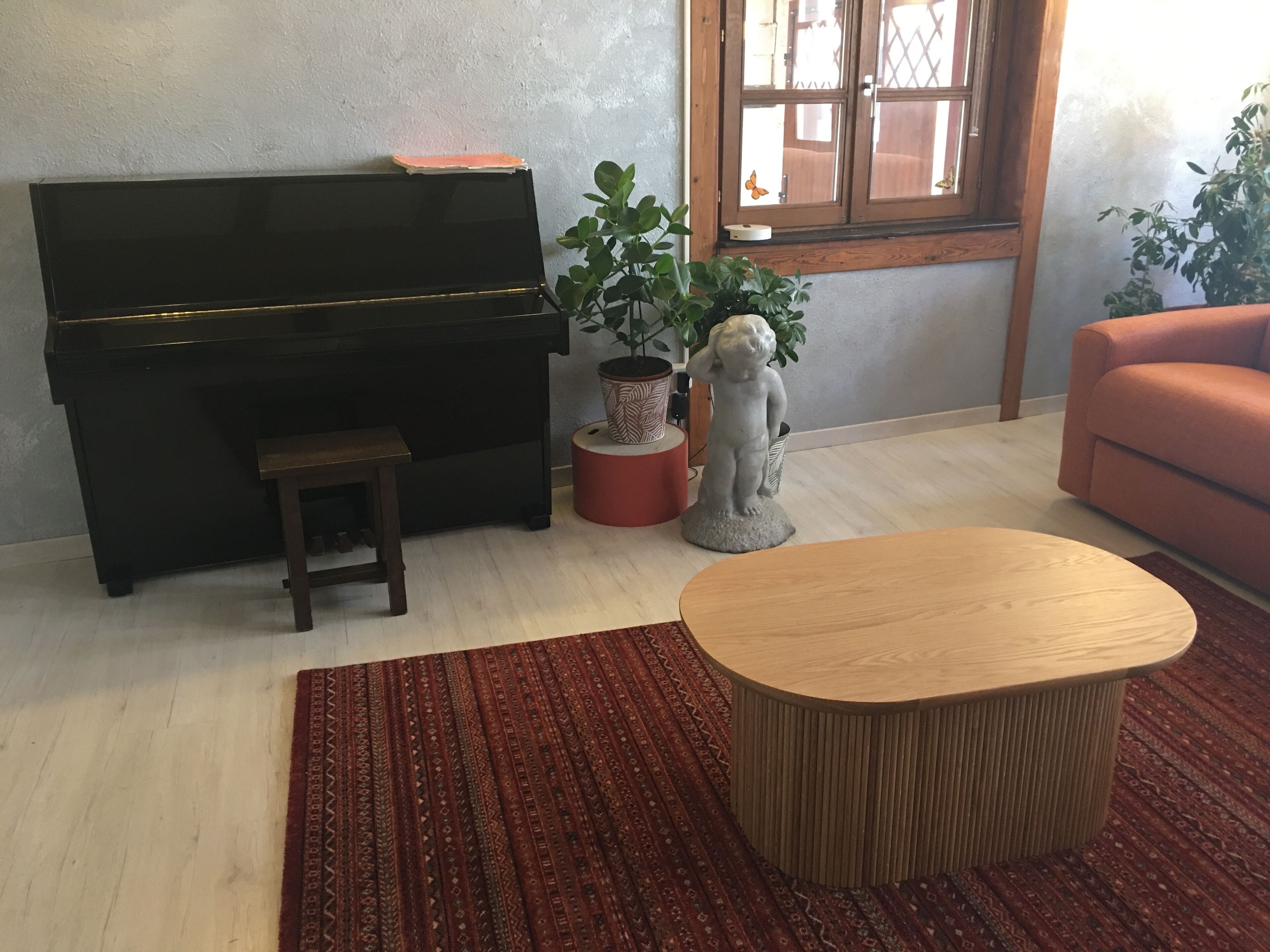 A living room with a black piano, a small stool, a wooden coffee table, a red patterned rug, and a statue of a child.