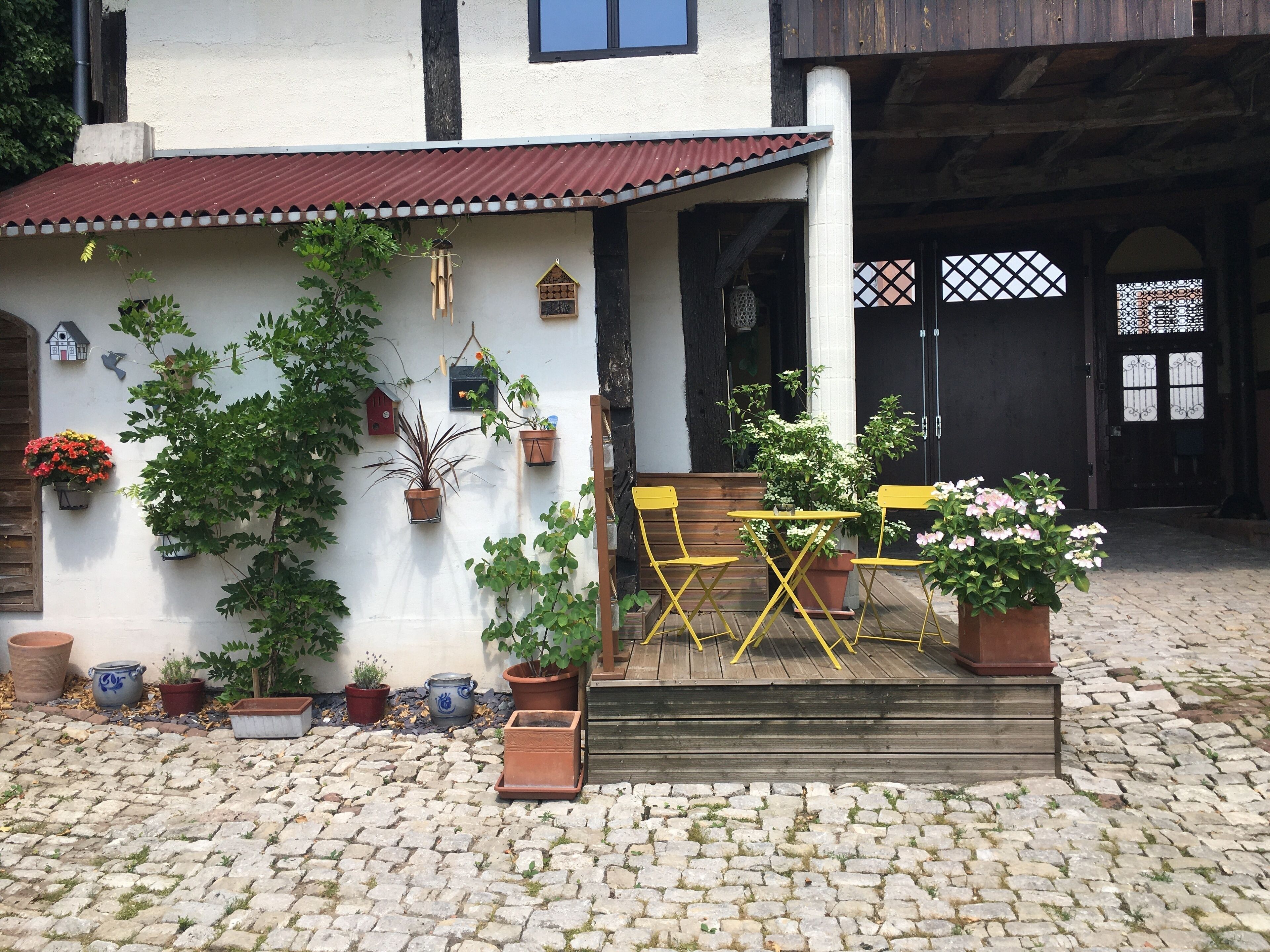 Un patio con mesa y sillas de madera, rodeado de plantas en macetas y un suelo de adoquines.
