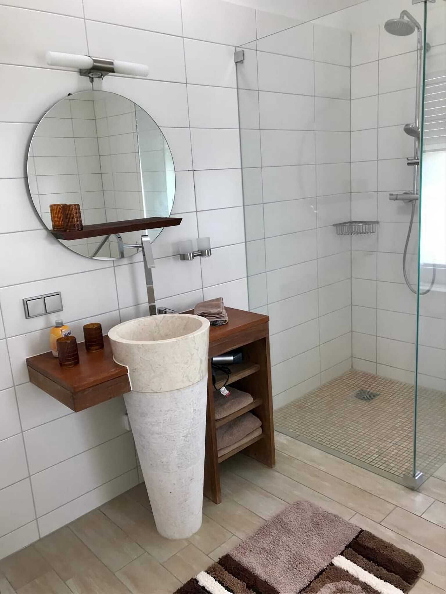 Un bagno moderno con un ampio specchio rotondo, un lavabo su piedistallo e una cabina doccia in vetro.