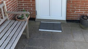 Una puerta blanca con un ventanal pequeño, un buzón, un banco y una planta en maceta.