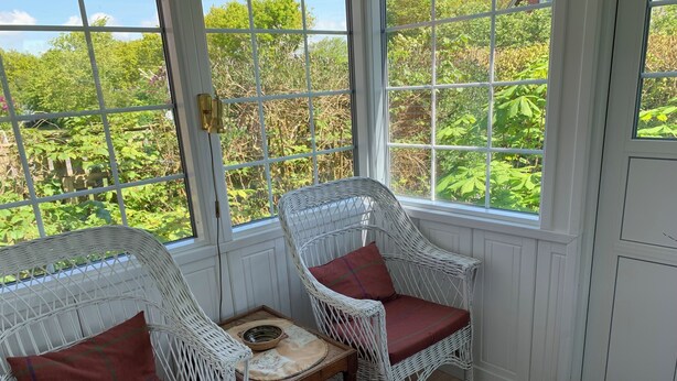 Un solárium con sillas de mimbre blanco y una pequeña mesa, con ventanas amplias que ofrecen vistas de abundante vegetación.