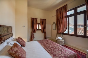 A hotel room with a bed, a chair, a window with curtains, and a view of buildings outside.