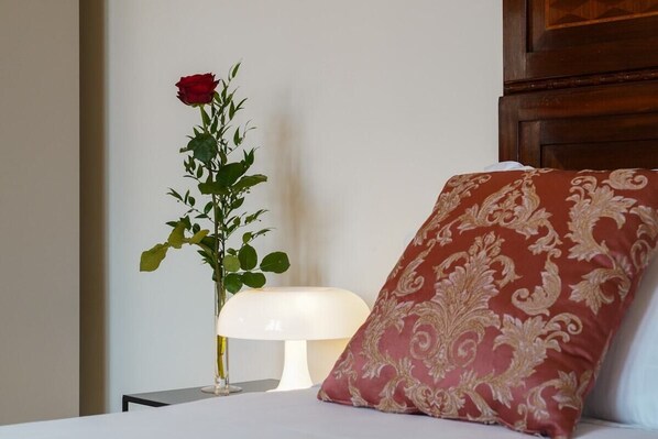 Een bed met een rood geborduurd kussen, een witte nachtkastje en een vaas met een enkele rode roos.