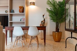 Een eetruimte met een houten tafel, twee witte stoelen, een grote potplant en een boekenplank met diverse spullen.