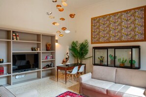 Una sala de estar con sofá, televisión, estanterías, mesa de comedor, sillas, una planta y una ventana con cactus.