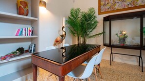 A dining area with a glass-top table, white chairs, a decorative bird sculpture, a potted plant, and a bookshelf with books and decorative items.
