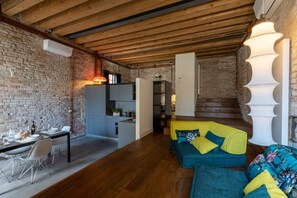 A modern interior with exposed brick walls, a wooden ceiling, a dining area with a table and chairs, and a sofa with colorful cushions.