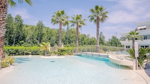 A clear blue pool surrounded by palm trees and greenery, with a bridge leading to a building.