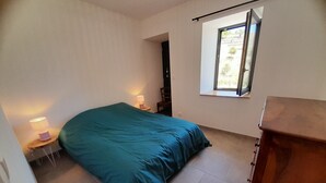 A bedroom with a bed, a nightstand with a lamp, a wooden dresser, and a window with a view of greenery. - Pretty house in southern Ardèche with view, in Cévennes hamlet (Lentillères)