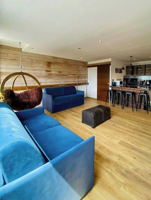 A modern living room with blue sofas, a hanging chair, and a kitchen area with bar stools.