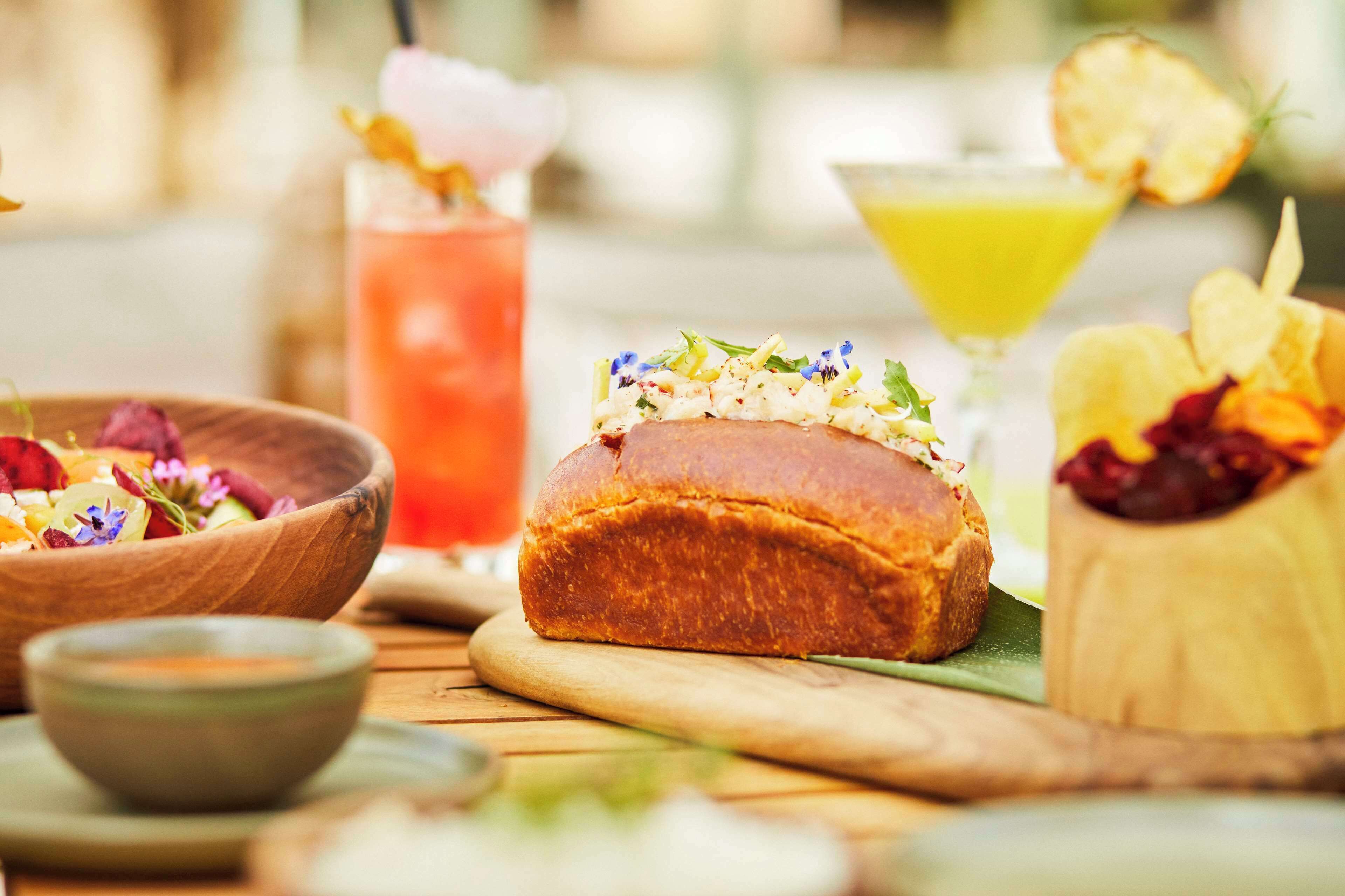 Una mesa de madera con un pan, un bol de ensalada, un cóctel y un recipiente pequeño con salsa.