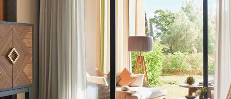Una habitación de hotel moderna con un ventanal grande, un sofá, una silla, una mesita y una lámpara decorativa.