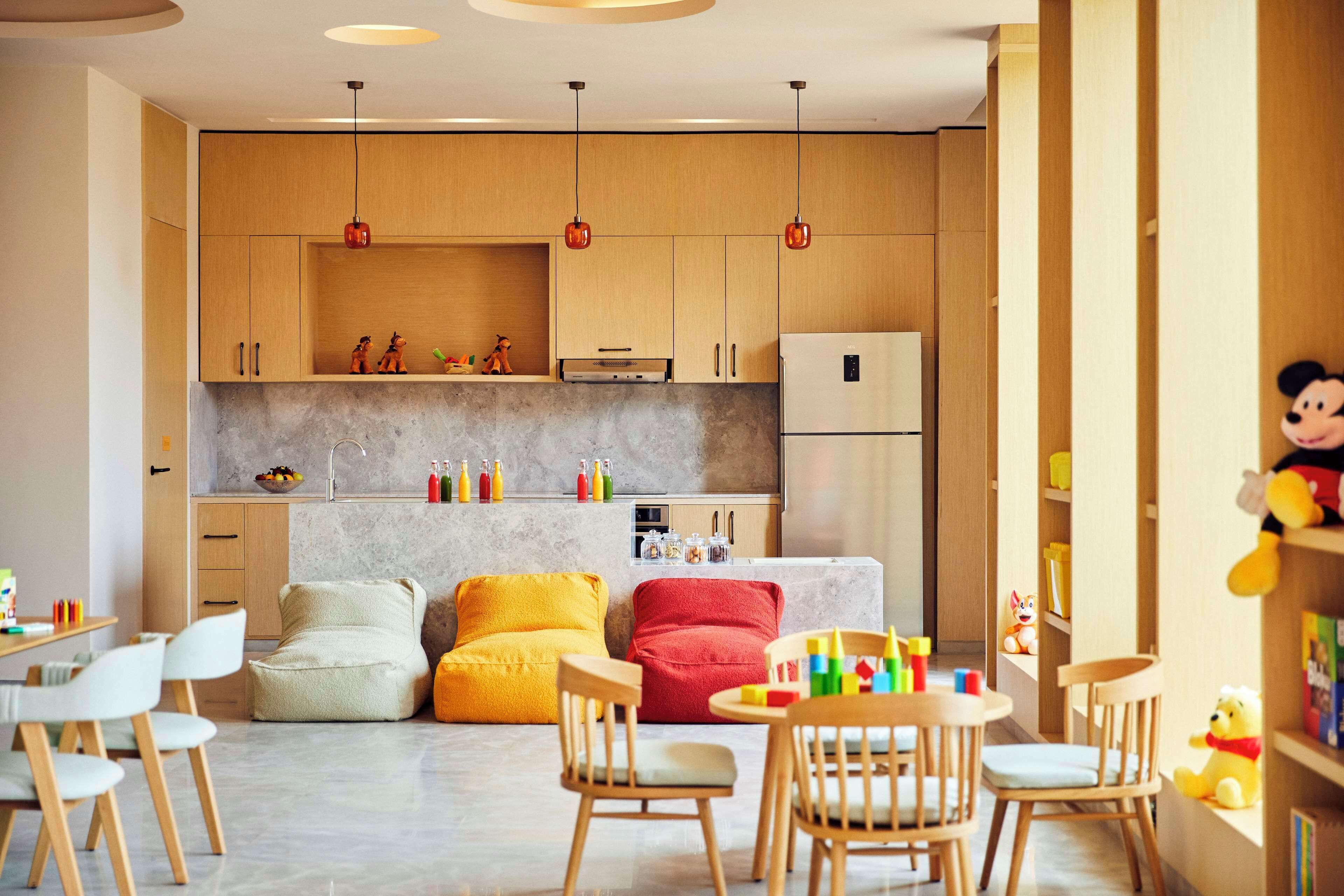 A modern kitchen with wooden cabinets, a refrigerator, and pendant lights. There are colorful pillows and a dining area with chairs.