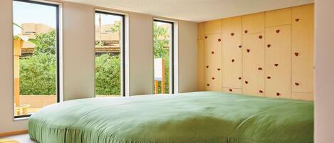 Un dormitorio moderno con una cama grande de color verde, una pared de madera con decoraciones en forma de corazón y ventanas amplias que ofrecen vistas al verdor exterior.