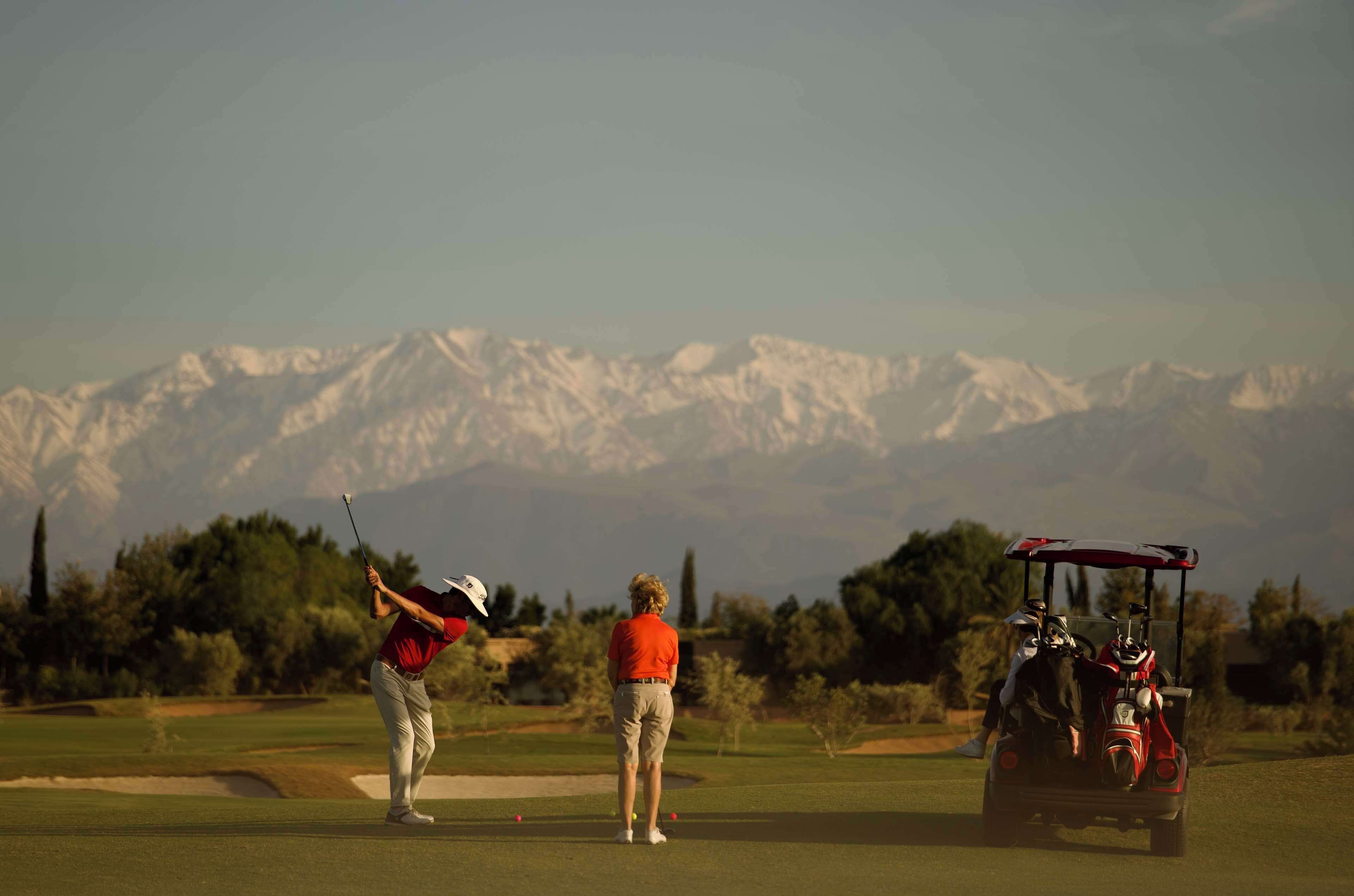 A golf course with a mountainous backdrop, a golf cart, and two people in red and white attire.