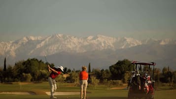 Un parcours de golf avec une chaîne de montagnes en arrière-plan, un chariot de golf et deux personnes vêtues de rouge et de blanc.
