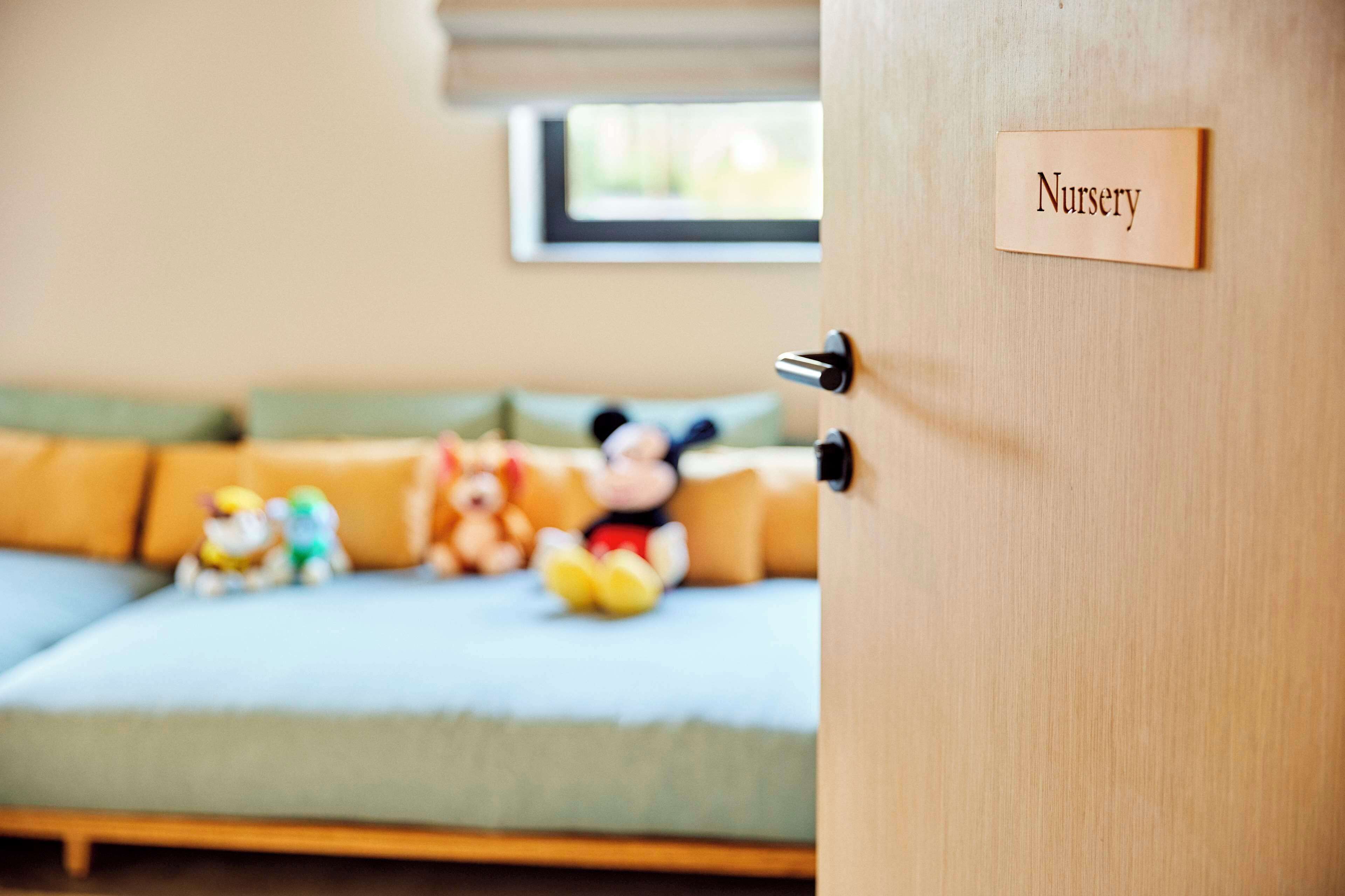 A room with a sign labeled "Nursery" on a door, a sofa with plush cushions, and a window in the background.