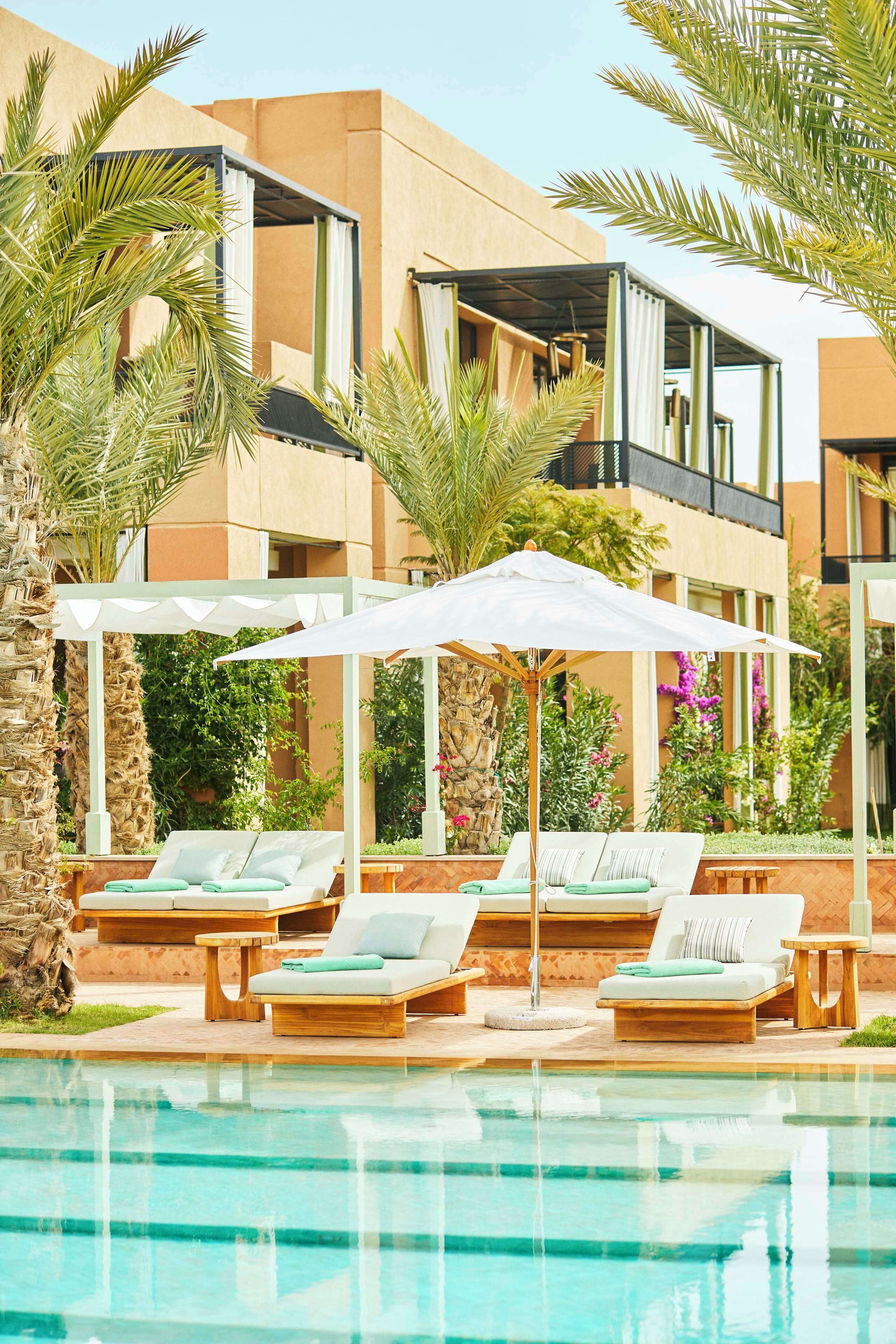 A pool area with lounge chairs and umbrellas, surrounded by palm trees and a building with a modern design.