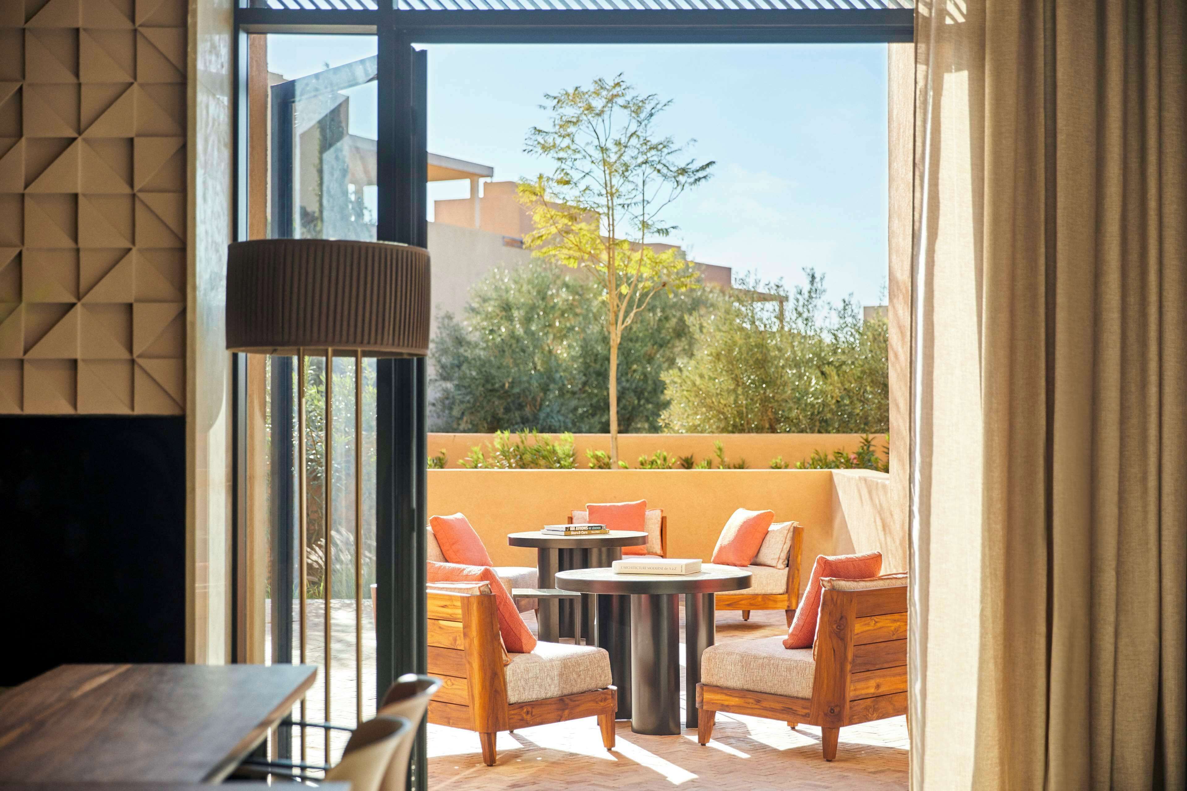 An outdoor seating area with wooden chairs and tables, a hanging light fixture, and a view of buildings and trees in the background.