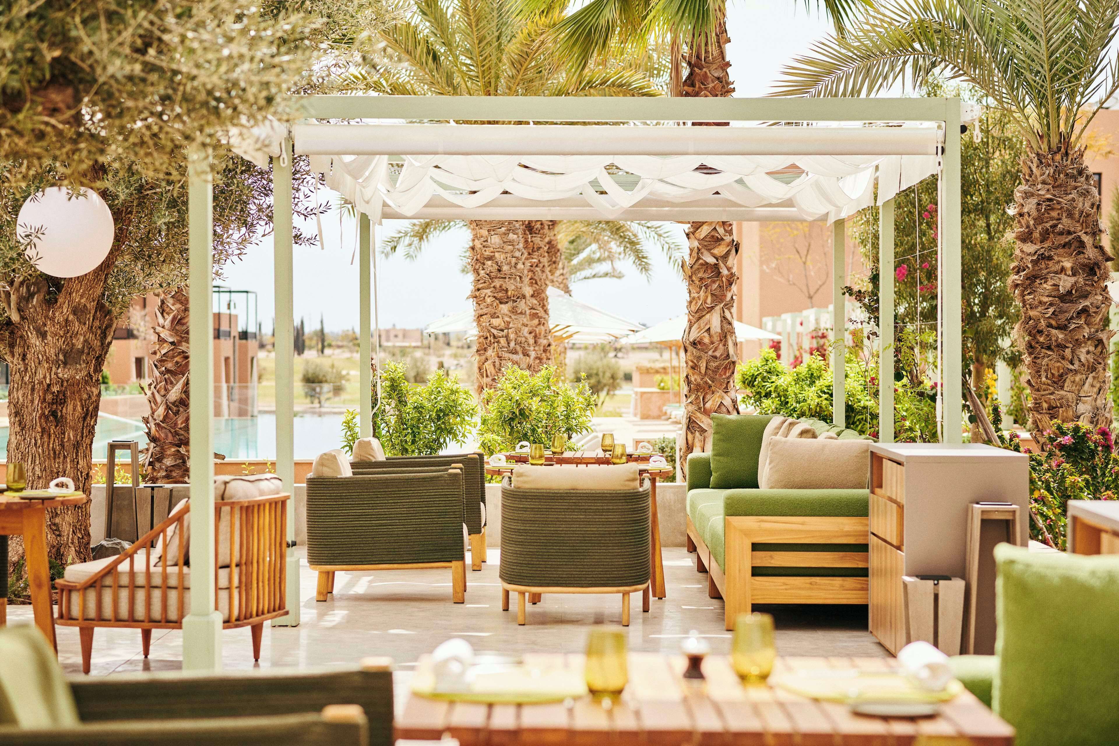 An outdoor seating area with a pergola, cushioned seating, and a pool in the background.