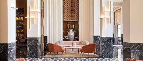 Un vestíbulo con suelo a cuadros, una zona de estar central con dos sillones y un sofá, un jarrón grande en un estante y un panel decorativo en la pared.