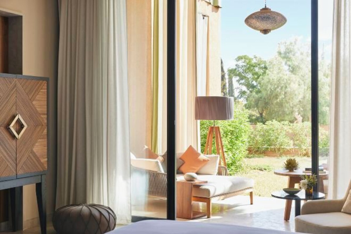 A modern hotel room with a large window, a sofa, a chair, a small table, and a decorative cabinet.