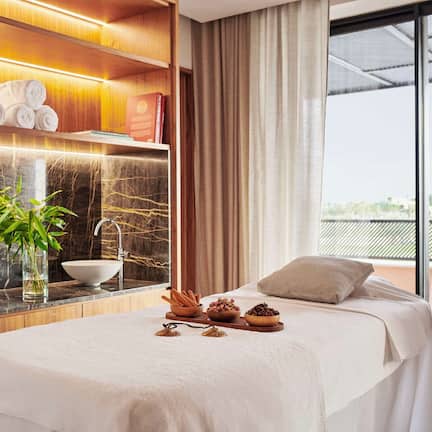 A hotel room with a bed, a wooden shelf with towels and a bowl, a plant, and a view of the outdoors.