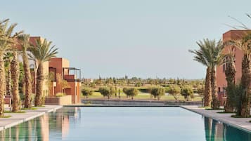 A modern hotel with a long swimming pool, palm trees, and a view of a golf course.