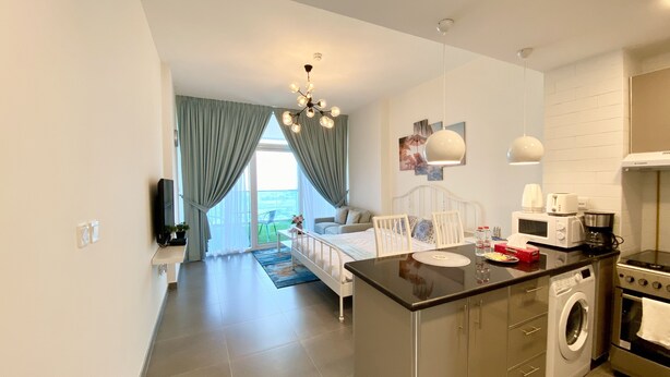 A modern kitchen with a central island, a washing machine, and a dining area with a view to a balcony. - Marco Polo - Pastel Studio with Stunning Aerial City Views (Dubai)