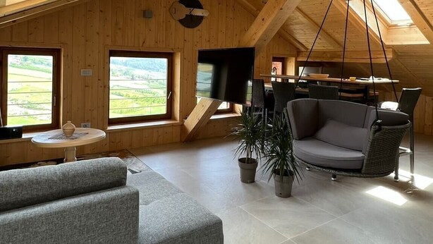 A spacious loft with a high ceiling, wooden beams, a large window, a flat-screen TV, a grey sofa, and a dining area.
