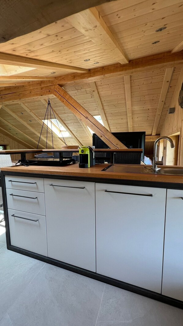 Una cocina en el ático con vigas de madera a la vista, una isla central con fregadero y un ventanal que proporciona luz natural.