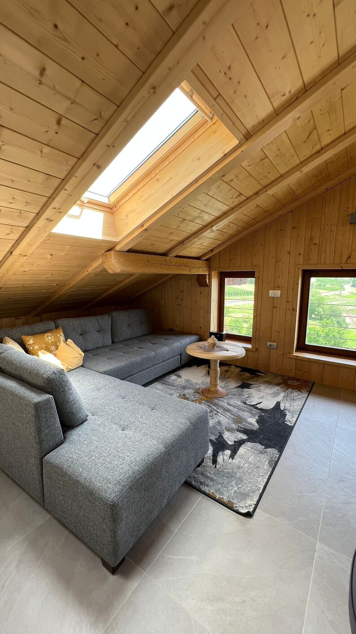 Ein Dachzimmer mit grauem Ecksofa, einem kleinen runden Tisch und Blick auf Grün.