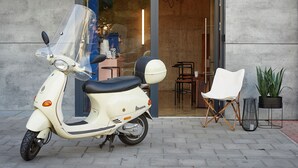 Una patineta blanca estacionada frente a un edificio con puerta de vidrio, una silla blanca y dos macetas.