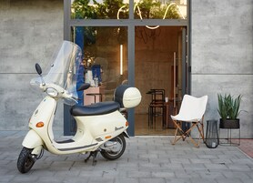A white scooter parked in front of a building with a glass door, a white chair, and two potted plants.