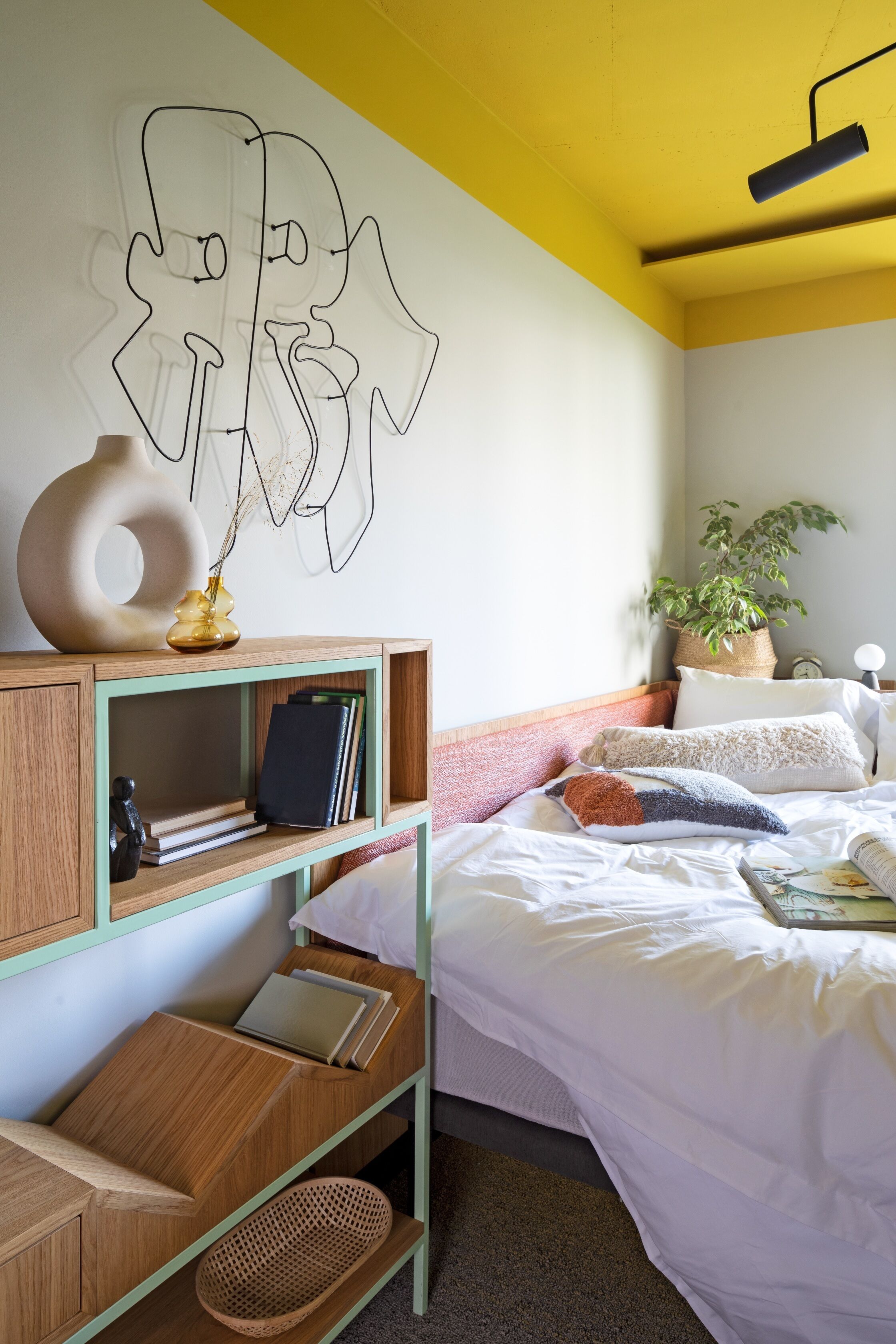 A bedroom with a bed, a wooden nightstand, a bedside table, a wall art piece, a potted plant, and a wall clock.