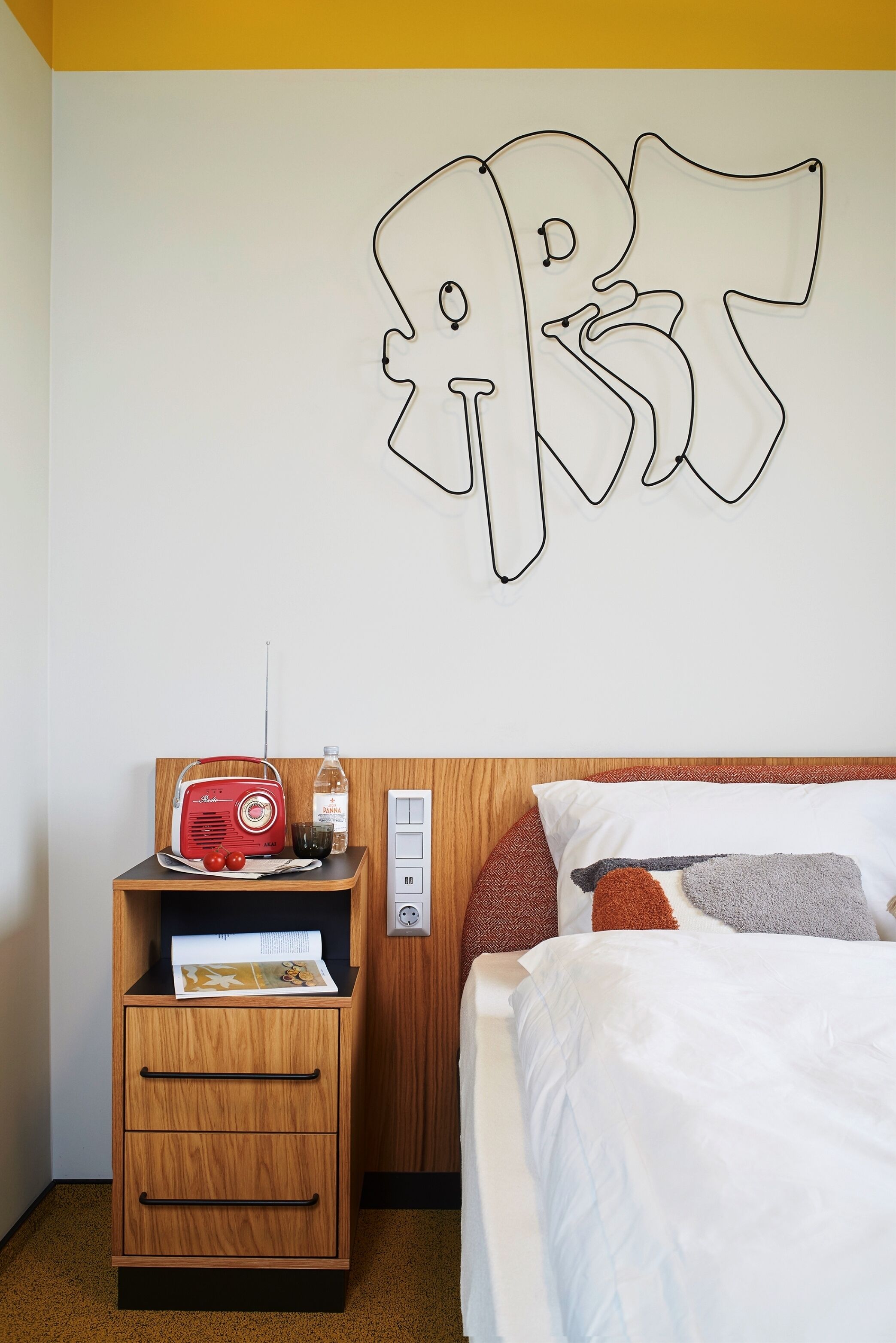 A hotel room with a wooden nightstand, a red radio, a bottle, and a magazine.